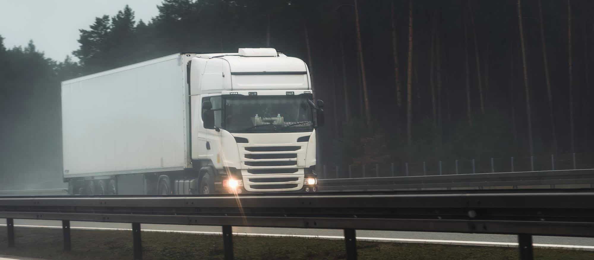 Hvit lastebil kjører på en våt motorvei med skog i bakgrunnen