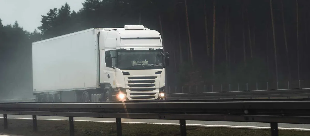 Hvit lastebil kjører på en våt motorvei med skog i bakgrunnen